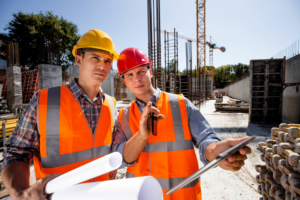 Zwei Bauleiter mit Helmen und Warnwesten besprechen Pläne auf einer Baustelle mit Kränen im Hintergrund.