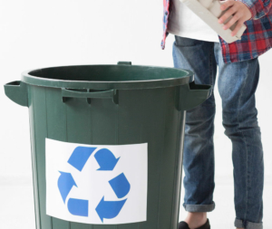 Person wirft Karton in grüne Recycling-Tonne mit blauem Recycling-Symbol.