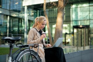 Frau sitzt mit Laptop und Kaffee vor modernem Bürogebäude neben ihrem Fahrrad.