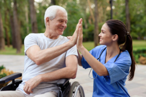 Pflegekraft gibt älterem Mann im Rollstuhl draußen im Park ein High-Five und lächelt.