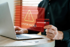 Warnmeldung „System Hacked“ erscheint auf Bildschirm, Person hält Kreditkarte vor Laptop in der Hand.